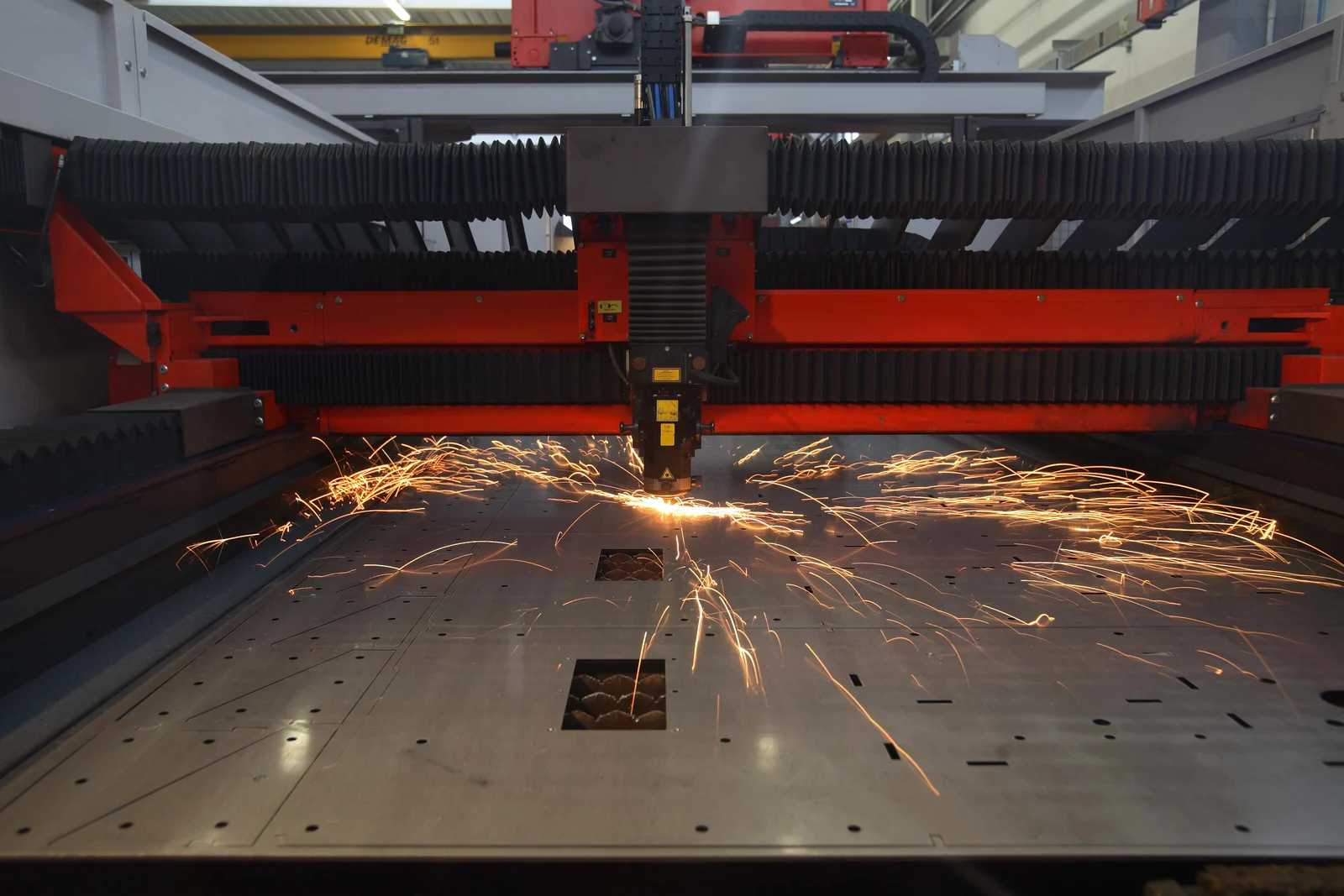 A laser cutting head actively cutting a metal sheet and throwing sparks inside a PTM Technology workshop.